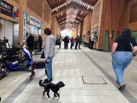 Owen Sennitt/BBC A woman with a black dog stands in the middle of Great Yarmouth's market while a woman wearing jeans and a black shirt walks past