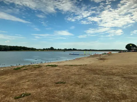 Alton Water is bordered by dried vegetation and dusty ground. There are green trees on the other side of the lake.