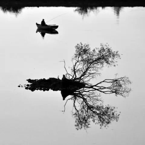 Victor Tregubov Silhouette of a fisher in a boat with a fishing road on a still loch in the evening, behind the silhouette of a small bit of land and branches protruding from the water.