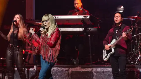 Redferns/Getty Images Anastacia singing on stage in front of a band. She has long blonde hair and glasses. She is wearing a red leather cowboy stale jacket with long red tassels along the arms. She is also wearing blue jeans and holding onto a microphone.