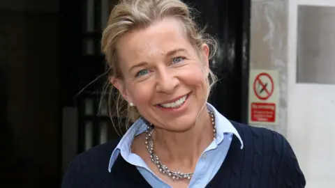 Katie Hopkins smiles at the camera with her head tilted to her right shoulder. She is wearing a navy blue jumper with a blue shirt underneath.