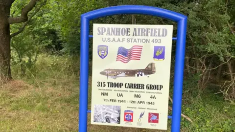 Qays Najm/BBC The sign for Spanhoe Airfield. The sing is held in a bright blue metal frame. The sign says "Spanhoe Airfield USAAF station 493". It has a picture of what looks like a Dakota aircraft, used during World War Two.