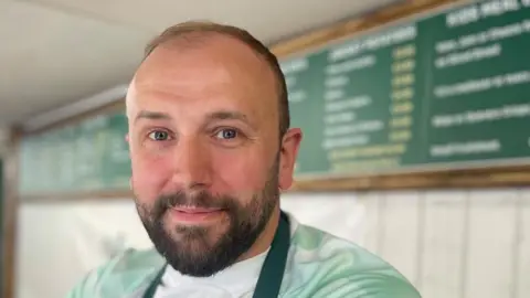 Simon Thake/BBC Dan Wilde, a bearded man in a green apron, stands before a board with a price list on it attached to the wall