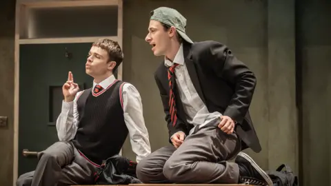 Marc Brenner Two boys sit on a table, wearing school uniform. They are looking out to the left of the photo. The are clearly on a stage.