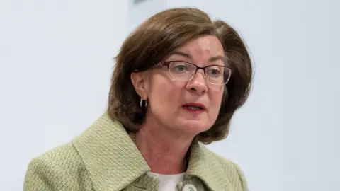 Senedd Cymru Eluned Morgan stood in the temporary Senedd debating chamber, wearing a green jacket and glasses. She has dark brown shoulder-length hair