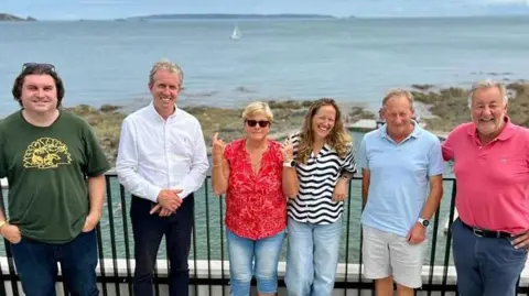 David Warr Six people stand in a line with their back to a railing in front of a bathing pool. The people are all smiling and looking towards the camera