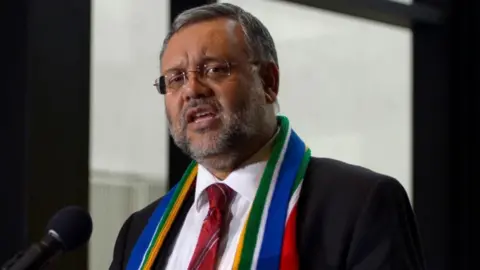 Ebrahim Rasool, wearing glasses and a dark jacket, with a scarf in the colours of South African flag