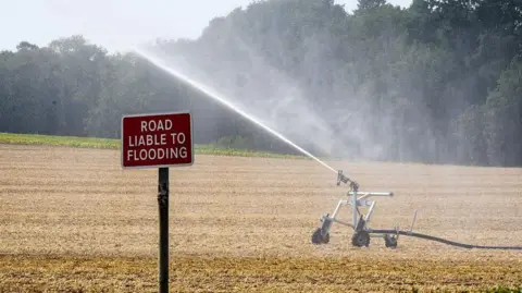 Getty Images A sprinkler system sprays a very dry field in Shropshire, with a sign visible that reads "Road liable to flooding".
