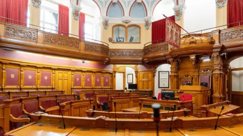 The empty States Assembly chamber. It has wood and leather benches and desks in concentric circles. There is an ornate wooden balcony with windows lined with curtains behind it. 