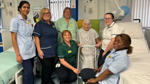 SaTH Gloria Malpass-Leek is sitting on a chair on a ward with a frame in front of her and six nursing staff in uniform sitting around her.