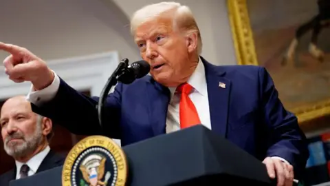 Getty Images US President Donald Trump, wearing a blue suit and orange tie, pointing at an audience member while he speaks at a lectern 