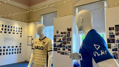 A photograph exhibition where photos have been stuck on to white walls. There are two mannequins wearing football shirts. 