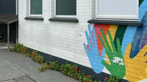 Paintworks NI An exterior wall of the Helping Hands building. It is painted white and there is a painted rainbow and colourful hands on it and three windows.