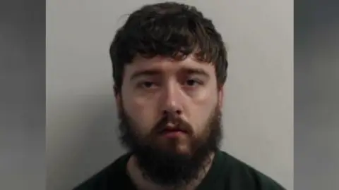 a police mugshot of a man with brown hair and a straggly brown beard who is wearing a dark-coloured round-necked top