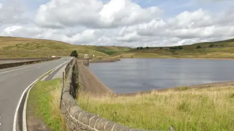 A wooden fence runs a long a road by a reservoir basin. Low green hills can be seen dotted with trees on the other side of the reservoir. 