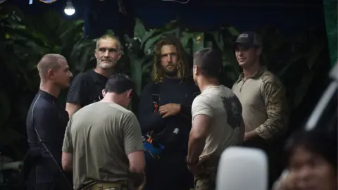 AFP/Getty A group of foreign divers and US soldiers, wearing light colored shirts, gather at the Tham Luang cave area as rescue operations continue for the 12 boys and their coach trapped at the cave in Khun Nam Nang Non Forest Park in the Mae Sai district of Chiang Rai province on July 6, 2018