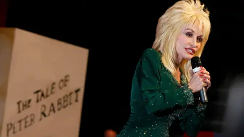 Dolly Parton stands holding a microphone wearing a green sequined dress in front of a beige sign reading 'The Tale of Peter Rabbit'. She has voluminous blonde hair and is wearing bright lipstick.