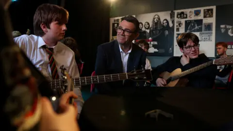 PA Media Prime Minister Andy Burnham speaks to young people during a visit to The Oh Yeah centre in Belfast. The young people are playing guitars.