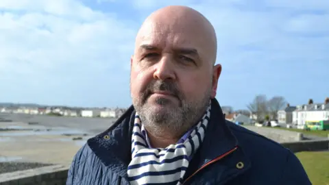 BBC Jonathan Le Tocq - A bald man with a black and grey beard, wearing a blue and white scarf and a blue jacket, standing in front of the Guernsey seafront at Bulwer Avenue with the beach on his right and the road on his left. 