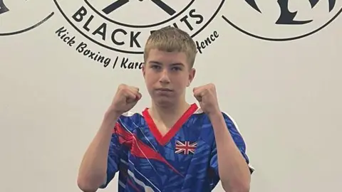 Supplied Lewis is wearing a blue, red and white kickboxing uniform. He is holding his fists up at the camera, in a pose. He has short blonde hair.