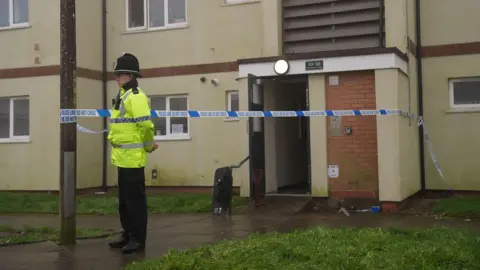 PA Media A police officer in a yellow high visibility jacket stands by a police cordon which is surrounding the entrance to a block of flats. There is police riot equipment visible by a doorway.