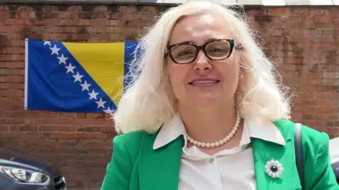 Lejla Golos is standing outside with the Bosnia-Herzegovina flag behind her, hanging from a wall. She has a Srebrenica flower pin badge on her green jacket, and she is wearing a white blouse with a pearl necklace. She has blonde hair and glasses, and has a handbag over her shoulder.