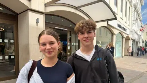Lily is on the left of the photo, wearing a grey hoodie and blue tshirt. She has brown hair. Alex is on the right with a black hoodie and white t-shirt. He also has brown hair. 