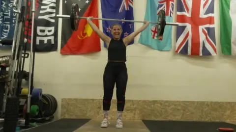 BBC Dawn pictured holding a large dumbbell over her head and smiling widely. She's wearing black leggings, a black sleeveless top and white and pink trainers, and has her blonde hair tied up in a ponytail. She's photographed indoors, inside her gym - which has cream walls and a brown wooden floor. Behind her are multiple flags, including the Union Flag, hung up on the wall. 