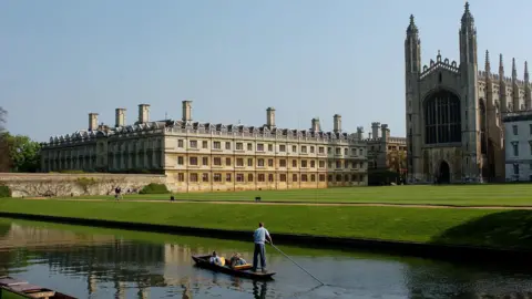 A river is in front of a large stretch of green grass that has large buildings which make up the University of Cambridge. A person is punting on the river.