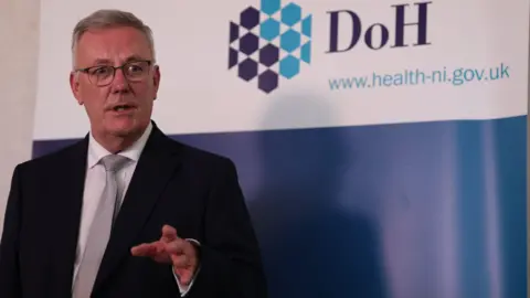 PA Media A man with white hair in a dark suit and glasses speaking in front of a white and blue DoH banner.