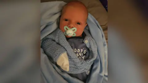 Family Baby Harley sucking a light blue dummy, wrapped in a blue fluffy blanket and baby grow.