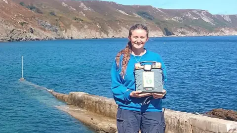 Tara McEvoy-Wilding Tara McEvoy-Wilding is standing beside a landing stage and azure blue sea. She is holding a blue pack containing a defibrillator and is wearing a blue top and darker blue shorts.