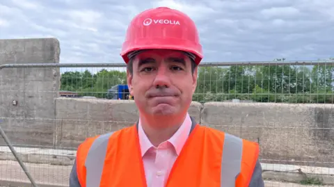 SAM READ/BBC A head and shoulders picture of Gavin Anderson who is looking at the camera with a slight smile. He is standing in front of a metal wire fence. Behind the fence a charred concrete wall can be seen and beyond that a tree line and a grey overcast sky. He is wearing a red hard hat and an orange high-visibility vest.