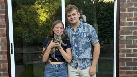 Grace Sangster & Ollie Vass Grace Sangster and Ollie Vass standing in front of patio doors holding a young tabby cat and smiling at the camera