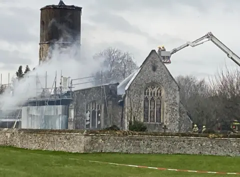 Jill Bennett/BBC The remains of the church with a fire service platform ladder hovering over it. One side of the building has scaffolding on it covered in metal hoardings. The roof has disappeared.