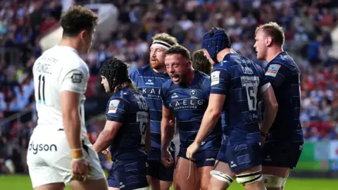 PA Media A group of Bristol Bears rugby players gather together to celebrate their matchwinning try against Bath at Ashton Gate. The Bristol players are wearing dark blue shirts and tops. There is a Bath player in an all-white kit in the foreground