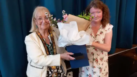 BBC Councillor Sue Eacock, left, pictured with Councillor Joanna Kane when she became the new Mayor of Redditch - Councillor Eacock is wearing a white jacket with a large gold chain around her neck. She is being handed flowers to mark the occasion.