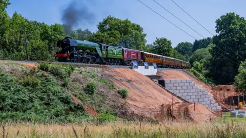 A green and red steam train going over a railway. The bank is brown with green foliage and there is a section which has grey bricks on it. 