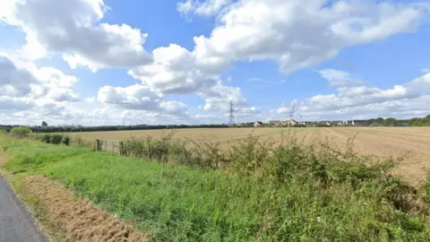 There is a landscape view of a Cirencester field beside a country lane. The outskirts of the field is bright green with grass and bushes, while inside the field the ground is dry and brown. On the nearest side there is a metal gate leading into the field, and on the opposite side there is a small group of houses. It is a bright sunny day with blue sky. 