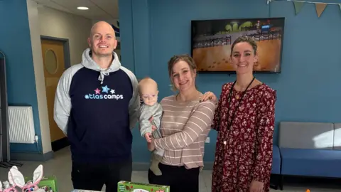Tzara Spurrier Three people smiling at the camera as they stand in a community centre hall. On the left is a tall man wearing an Atlas Camps hooded jumper. In the middle is Tzara Spurrier, who is holding her baby in her arms. On the right is another woman who is wearing a red long-sleeved floral dress and has a whistle around her neck.