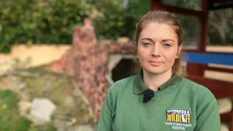 A woman in a branded green sweatshirt stands in front of an animal enclosure at a wildlife park