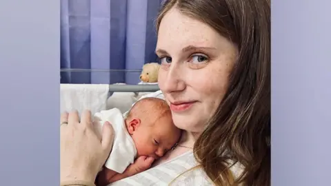 A smiling woman with brown hair cuddles a newborn baby on her chest. She is wearing a white and grey striped top, with the baby in a white grow. A blue curtain hangs in the background of the hospital setting. 