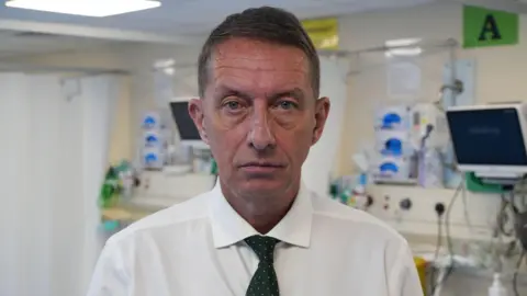 John Fairhall/BBC Matthew Hopkins standing in a hospital surgery room, wearing a white shirt and a black tie spotted with white specks. He has blue eyes and is staring directly into the camera. He appears to be aged at least in his 50s.