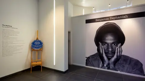 David Rowan/Birmingham Museums Trust A black and white photo of Benjamin Zephaniah, which fills an entire wall, is next to a blue plaque resting on a wooden stand