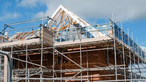 A stock image showing a house being built, it has red brick walls and scaffolding around the top part leading to a pointed roof made only of the timbers. The sky behind it is blue with some cloud.