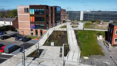 A drone photo of the top of the boulevard, showing two flights of steps with newly planted trees and patches of grass, with cars and buildings on each side and Taunton station at the end.