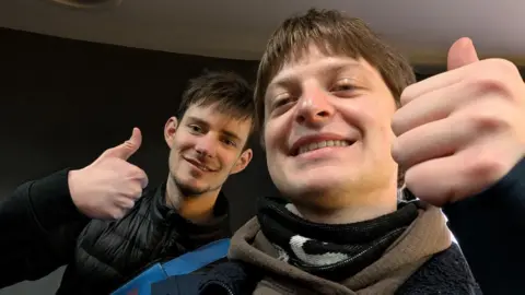 Handout A selfie, featuring close-ups of two young men, smiling. Both have short brown hair and are giving "thumbs up" gestures. The man on the left is wearing a black jacket and has a blue bag strap over his shoulder. 