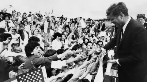 A black and white archive image of the then US President John F Kennedy meeting some very excited well-wishers at New Ross, County Wexford in 1963. He has short, fair hair and is wearing a dark suit. Many of the fans are young women and girls. Some are waving US flags and are stretching out to shake hands with Kennedy. 