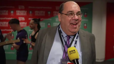 Mark Stupple gives a TV interview inside a room at Wembley stadium with players behind him. He is bald, with brown hair at the sides, and wears glasses and a suit and tie.