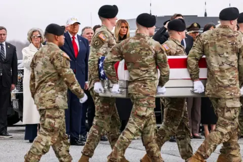 Getty Images Members of a US Army carry team transports a flag-draped transfer case containing the remains of Sergeant First Class Nicole M Amor during a dignified transfer at Dover Air Force Base in Dover, Delaware. In the background, President Donald Trump, wearing a dark blue suit, red tie and white USA baseball cap, is saluting. First Lady Melania Trump is next to him.